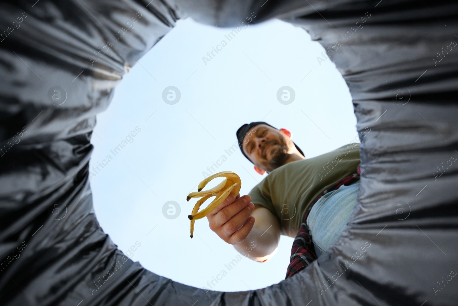 Photo of Man throwing banana peel into garbage bin outdoors, bottom view