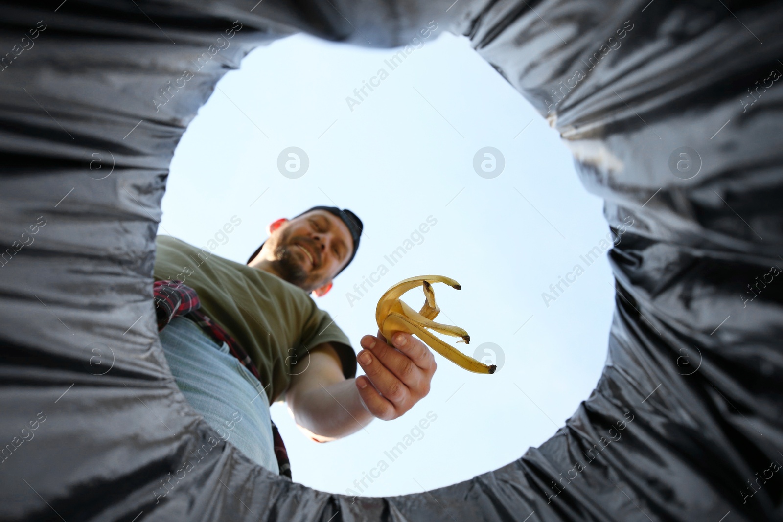 Photo of Man throwing banana peel into garbage bin outdoors, bottom view