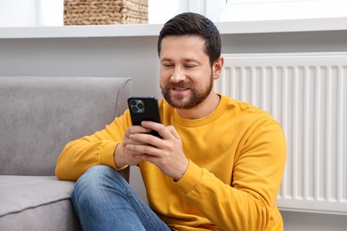 Happy man using smartphone near heating radiator at home