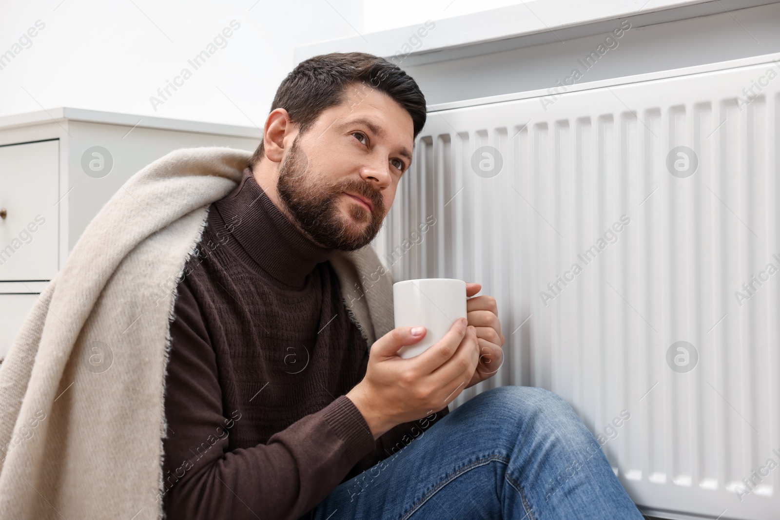 Photo of Man with cup of hot drink near heating radiator at home