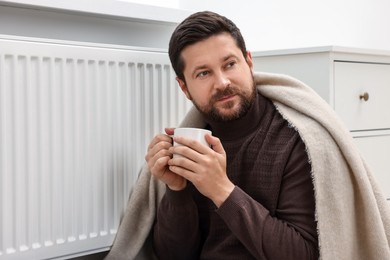 Man with cup of hot drink near heating radiator at home