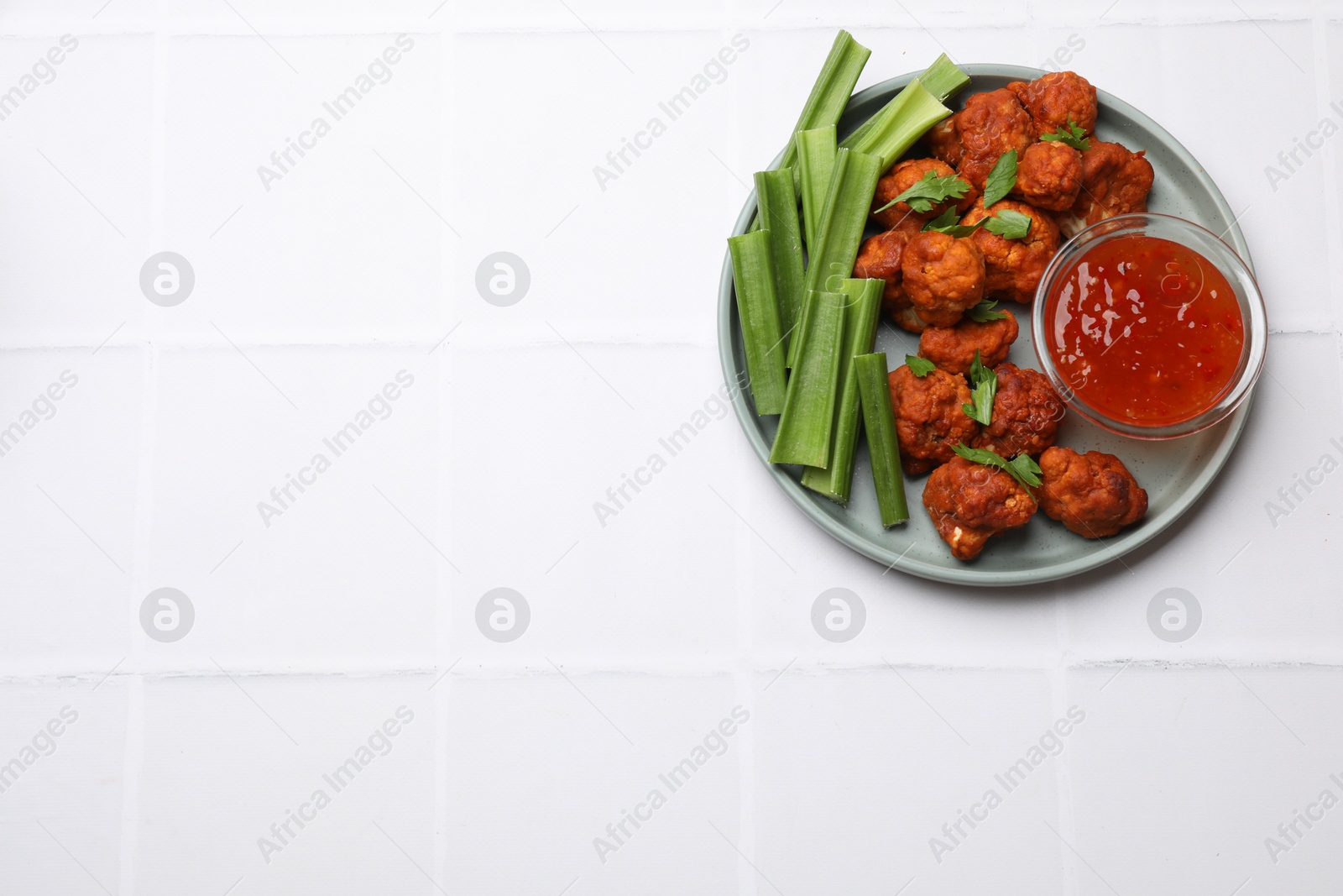 Photo of Baked cauliflower buffalo wings with celery and sauce on white tiled table, top view. Space for text