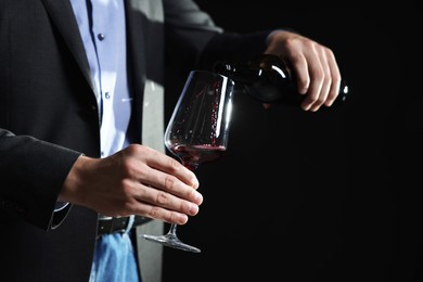 Photo of Man pouring red wine from bottle into glass on black background, closeup. Space for text