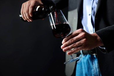 Photo of Man pouring red wine from bottle into glass on black background, closeup. Space for text