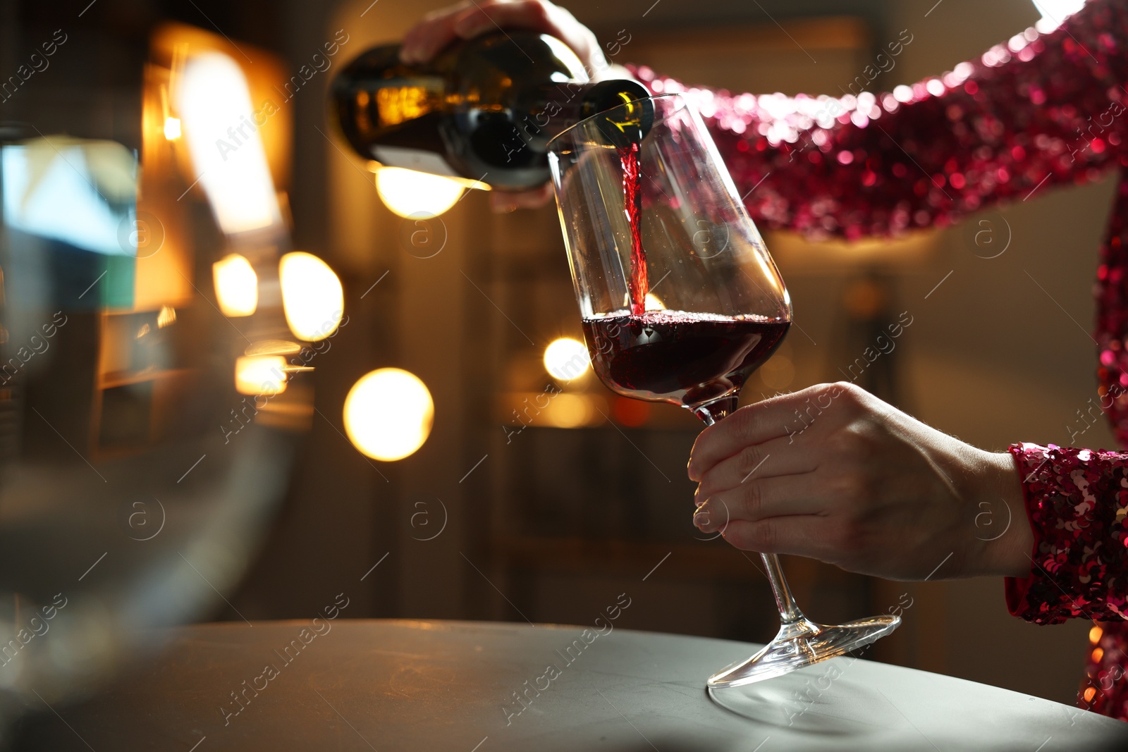 Photo of Woman pouring red wine from bottle into glass at table indoors, closeup