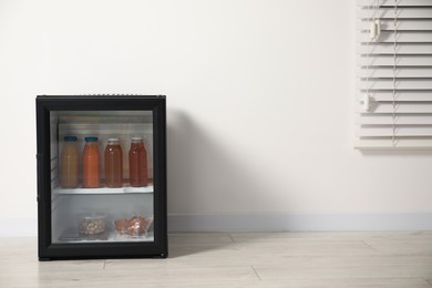 Mini refrigerator with different drinks and snacks near white wall, space for text