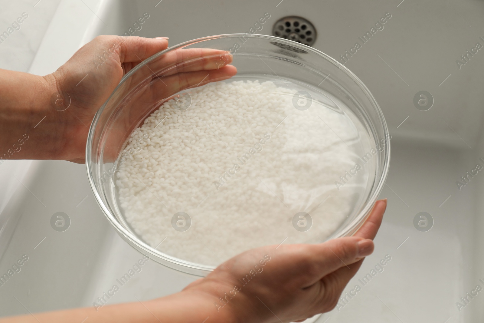 Photo of Woman holding bowl with rice and water above sink, closeup