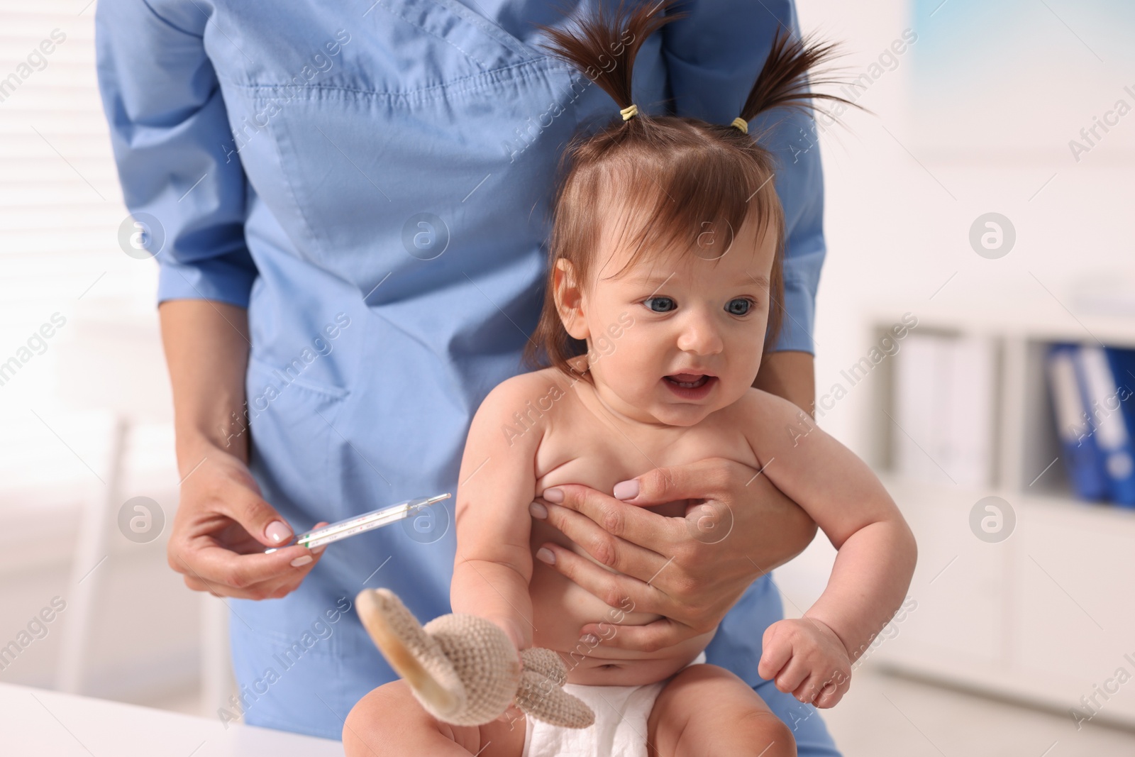 Photo of Pediatrician examining cute little girl with thermometer in clinic, closeup. Checking baby's health