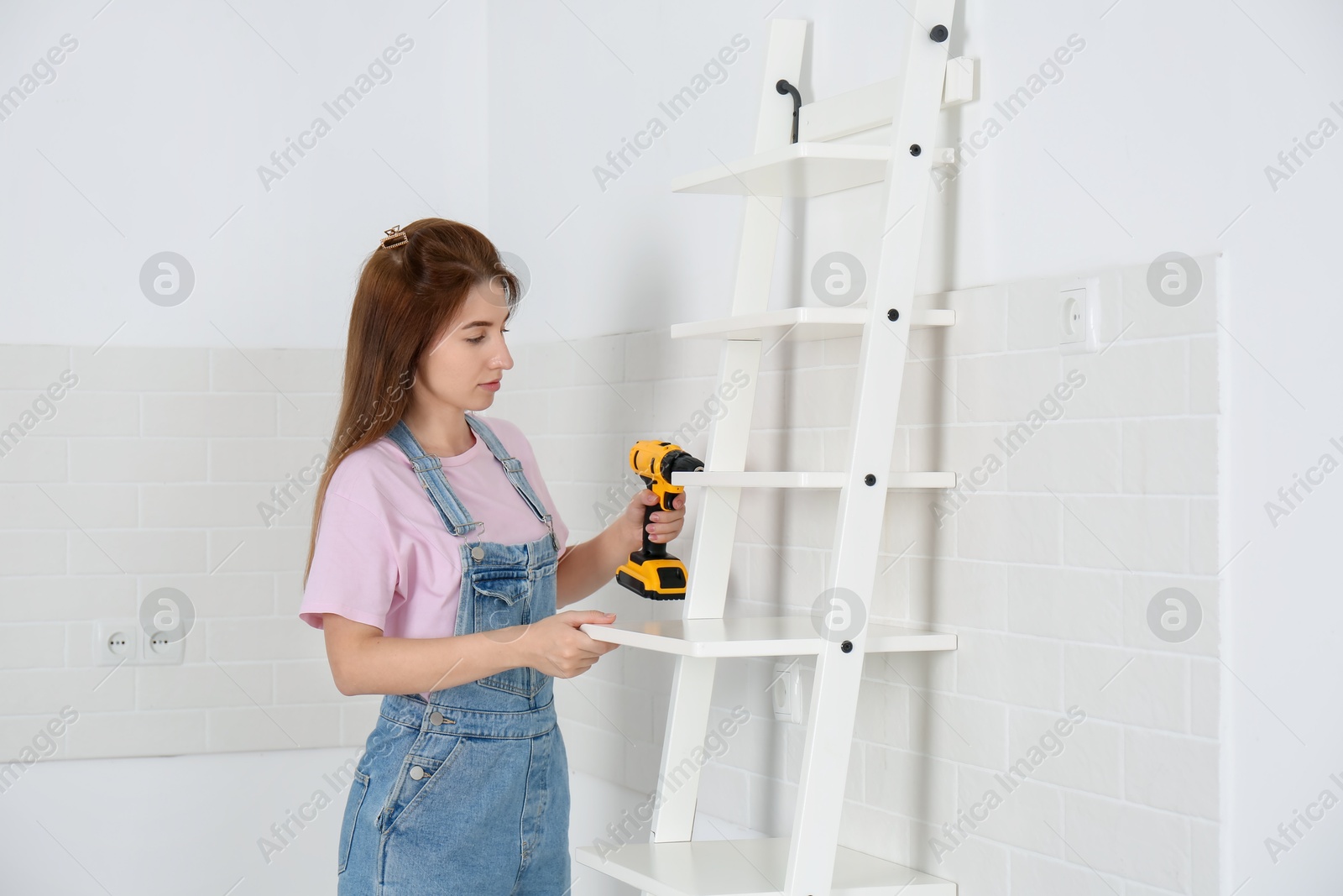 Photo of Woman with electric screwdriver assembling ladder shelf indoors