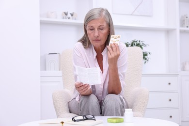 Senior woman with pills reading medicine instruction in armchair at home