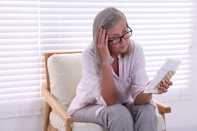 Senior woman with pills reading medicine instruction in armchair at home