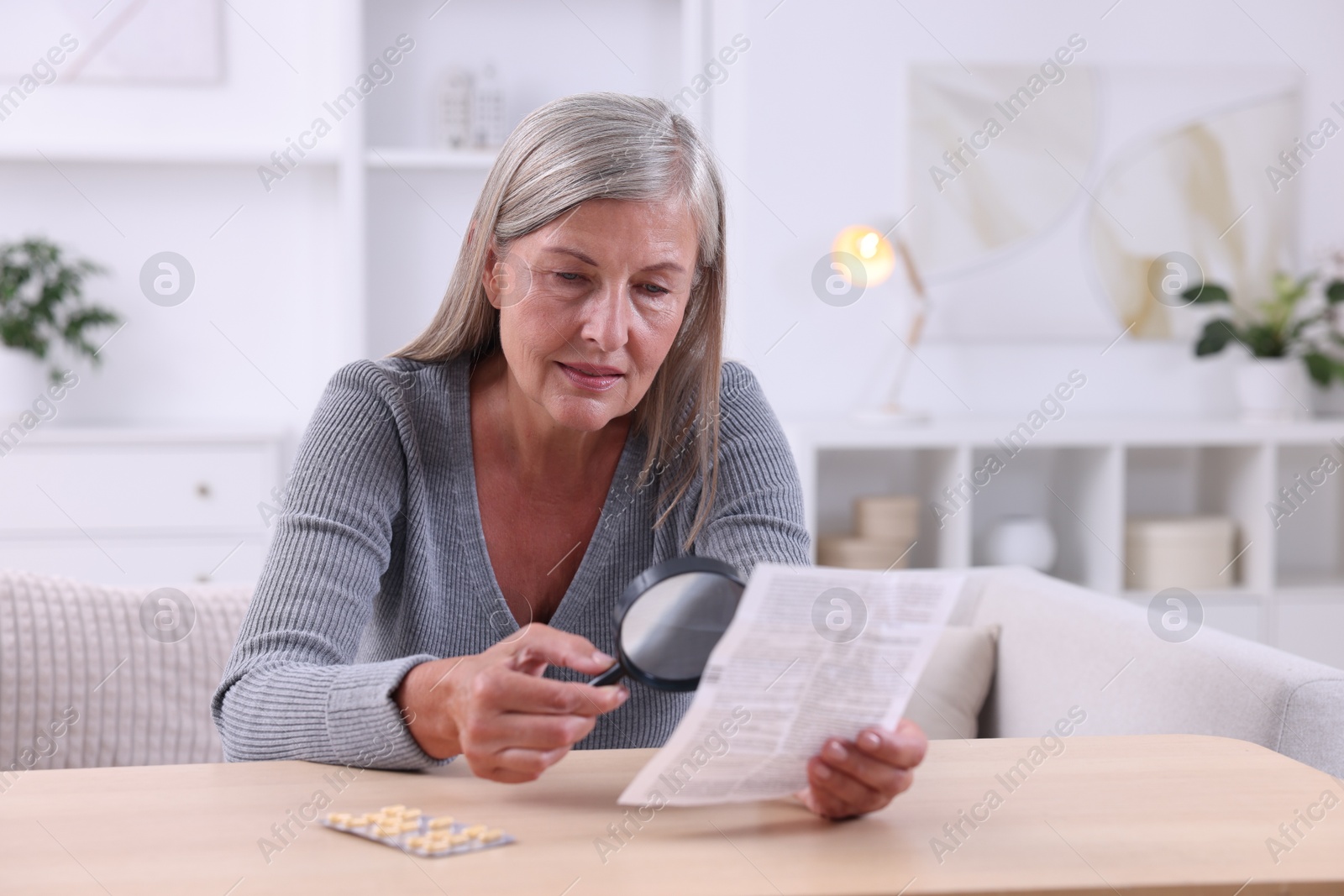 Photo of Senior woman with magnifying glass reading medicine instruction at table indoors