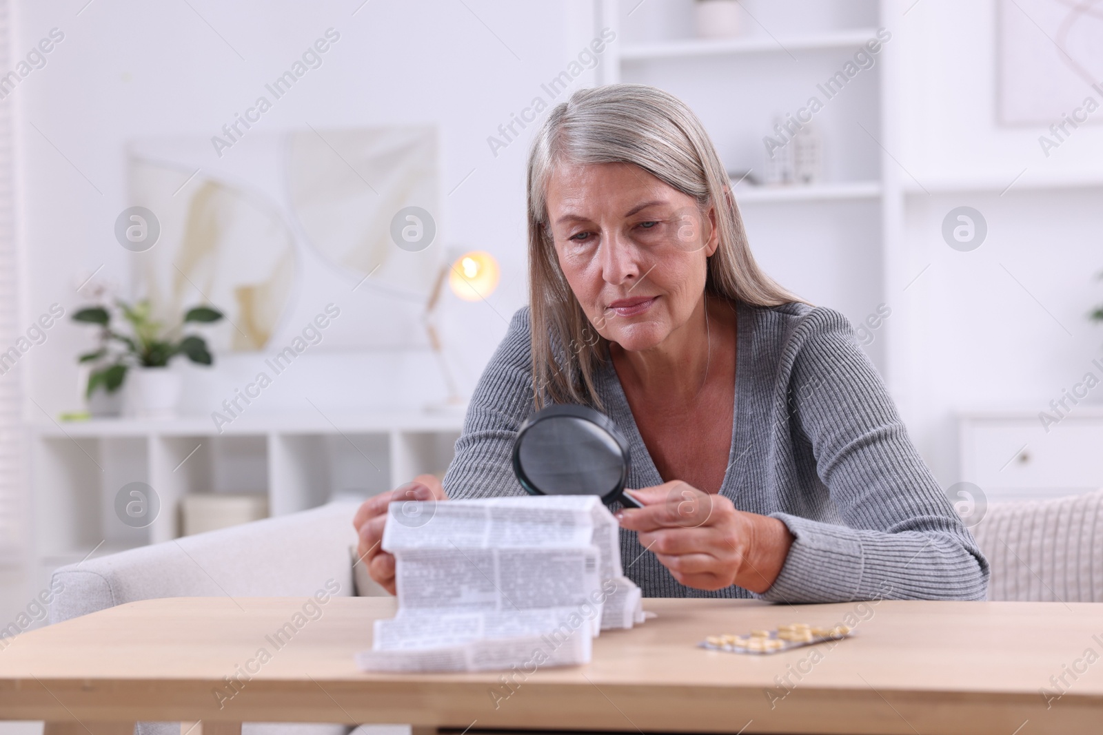 Photo of Senior woman with magnifying glass reading medicine instruction at table indoors