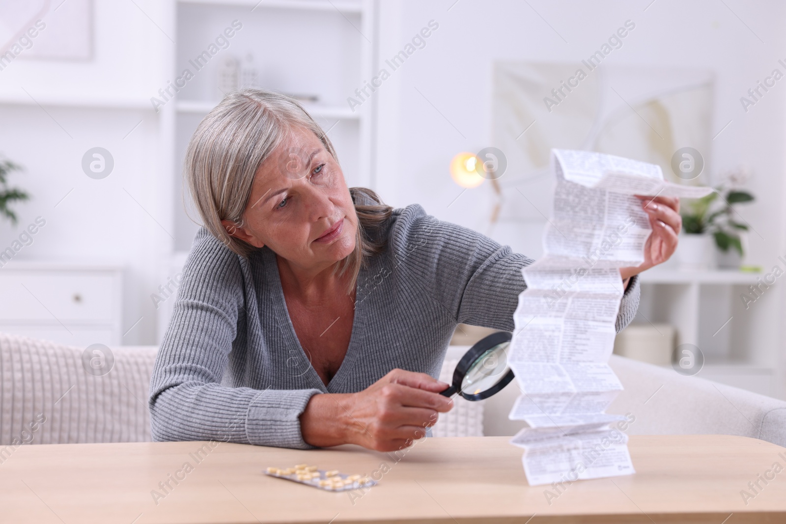 Photo of Senior woman with magnifying glass reading medicine instruction at table indoors