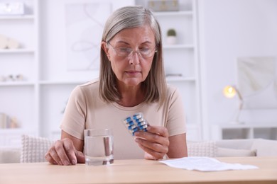 Photo of Senior woman with pills and glass of water at table indoors