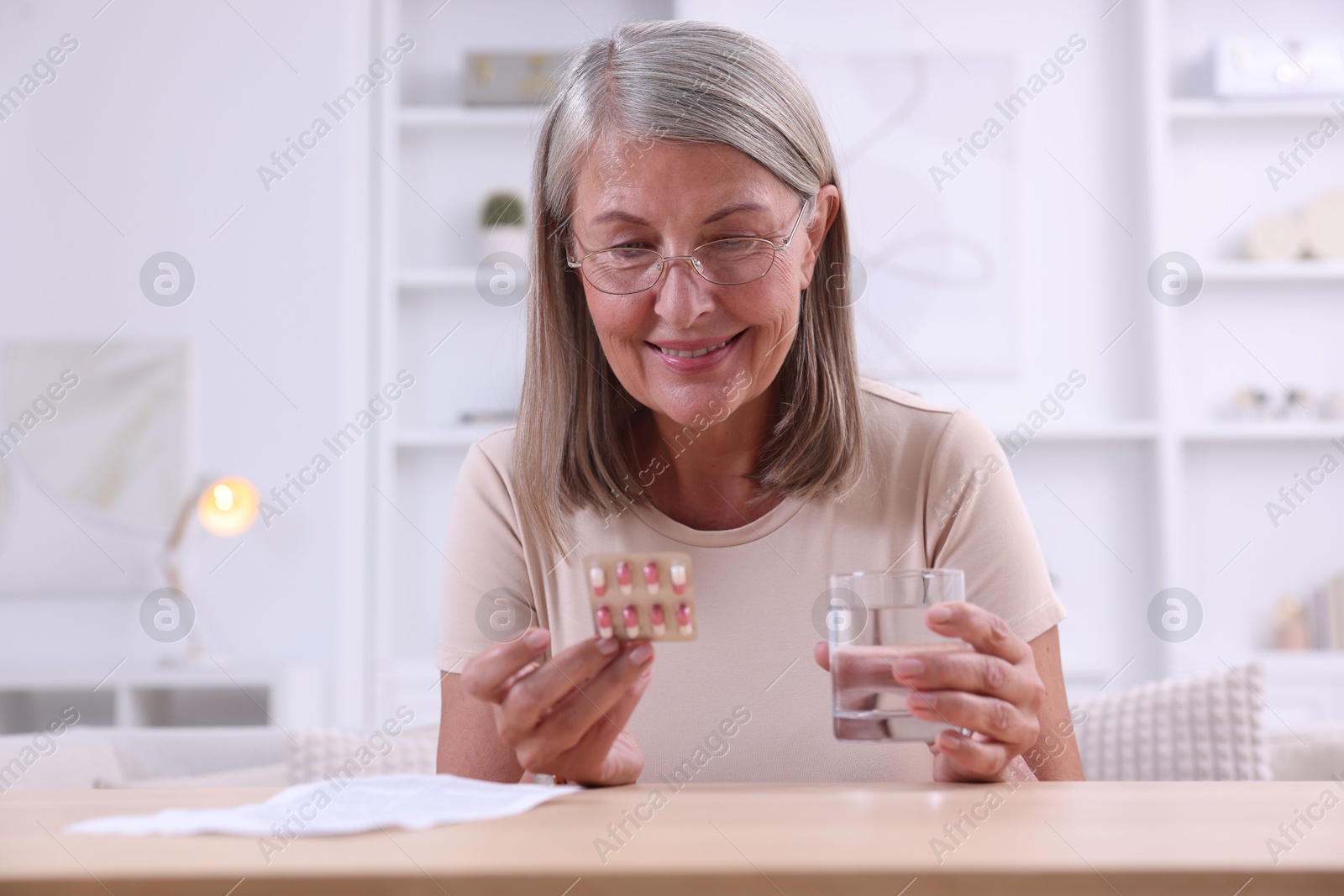 Photo of Senior woman with pills and glass of water at table indoors