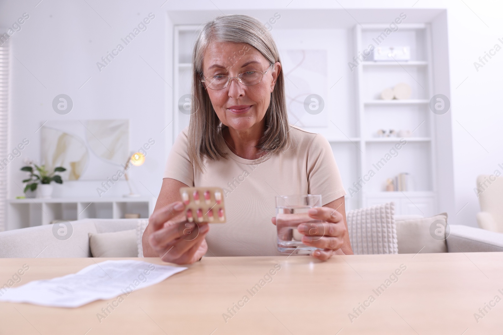 Photo of Senior woman with pills and glass of water at table indoors