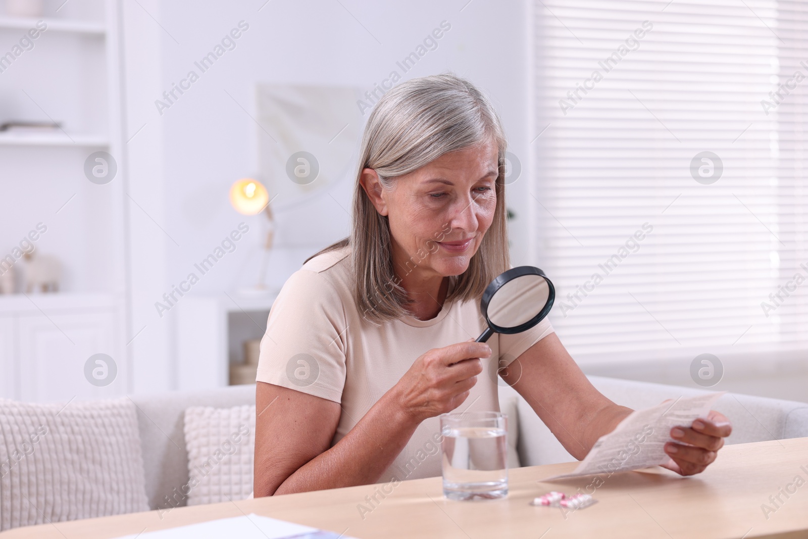 Photo of Senior woman with magnifying glass reading medicine instruction at table indoors