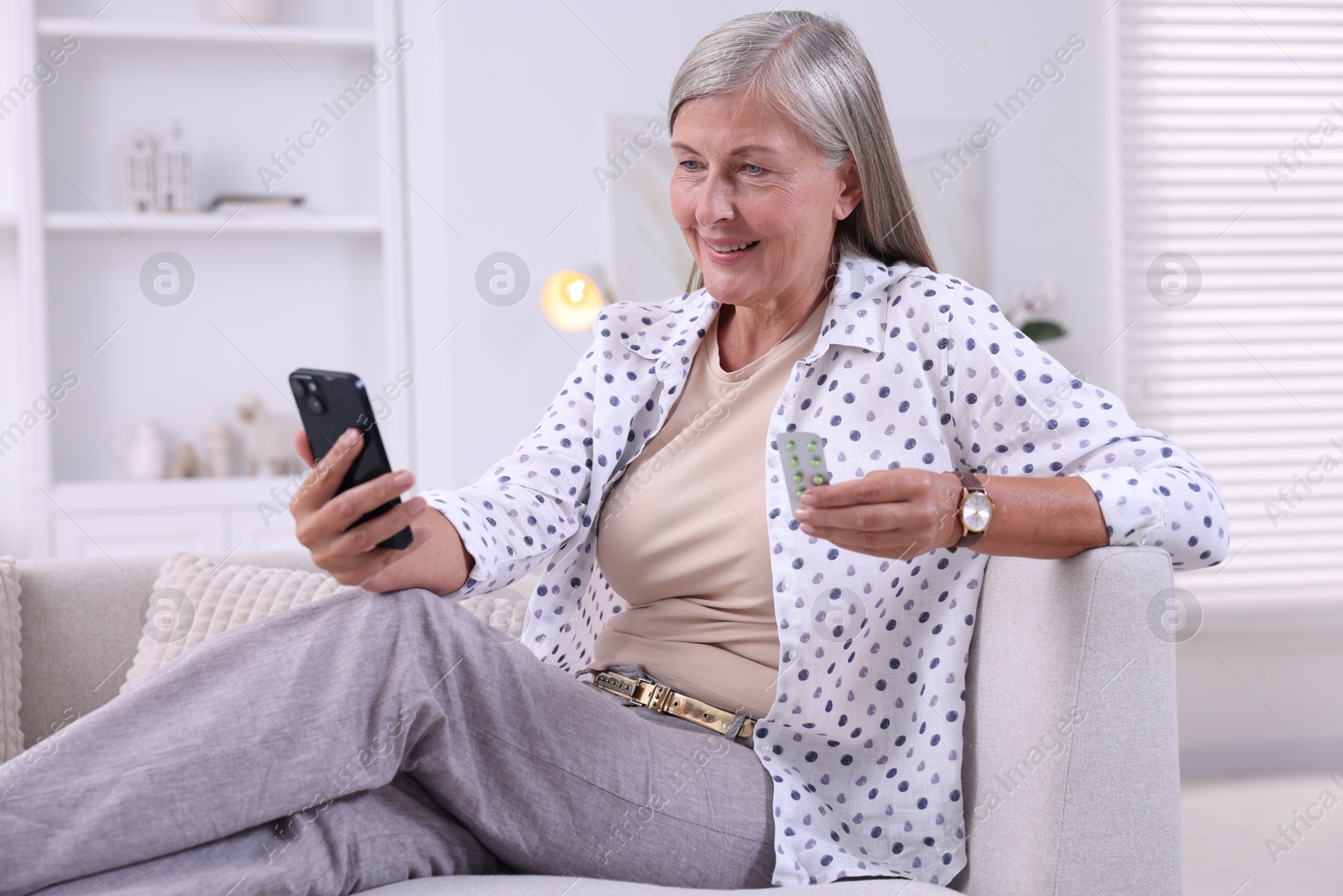 Photo of Senior woman with pills using smartphone on sofa at home