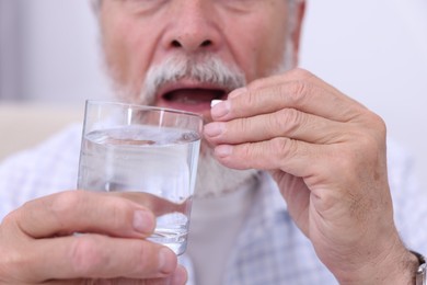 Senior man with glass of water taking pill at home, closeup