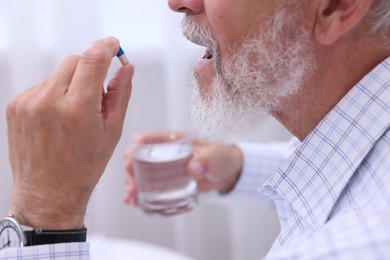 Senior man with glass of water taking pill at home, closeup