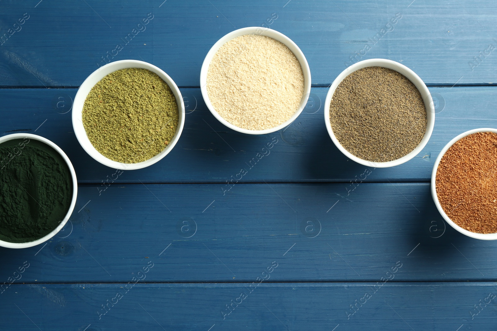 Photo of Different superfood powders in bowls on blue wooden table, flat lay