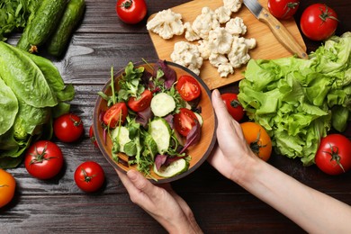 Photo of Healthy vegetarian food. Woman holding bowl of salad at wooden table with vegetables, top view