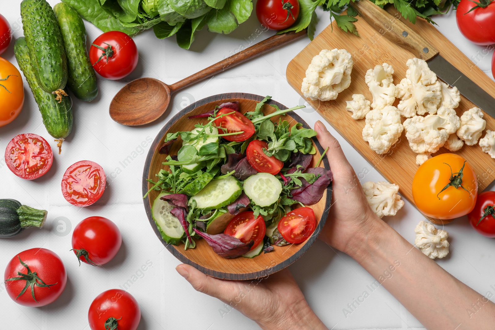 Photo of Healthy vegetarian food. Woman holding bowl of salad at white tiled table with vegetables, top view