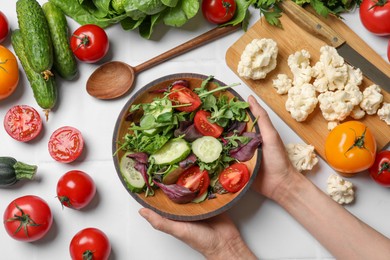 Healthy vegetarian food. Woman holding bowl of salad at white tiled table with vegetables, top view