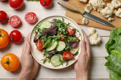 Photo of Healthy vegetarian food. Woman holding plate of salad at light wooden table with vegetables, top view