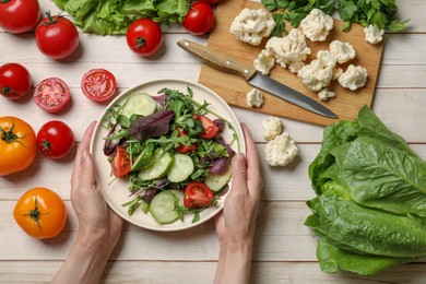 Healthy vegetarian food. Woman holding plate of salad at light wooden table with vegetables, top view