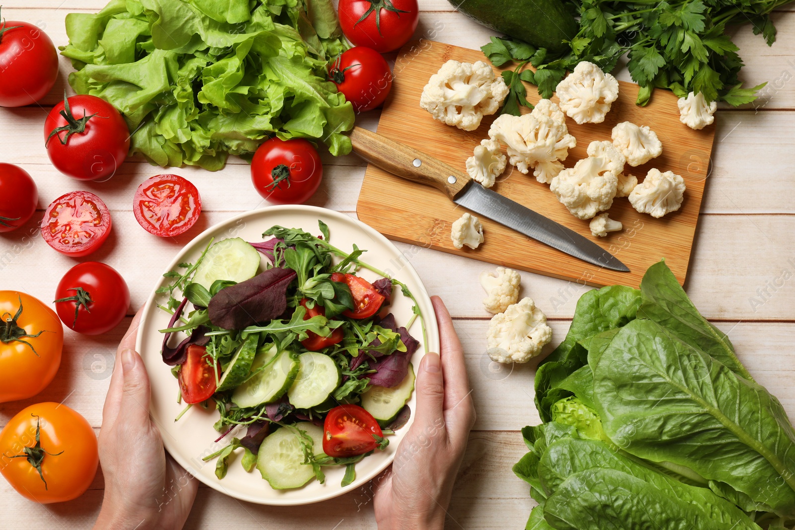 Photo of Healthy vegetarian food. Woman holding bowl of salad at light wooden table with vegetables, top view