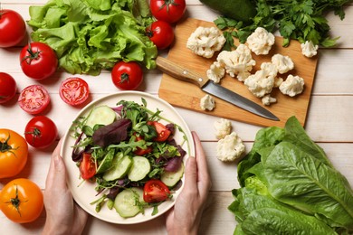 Photo of Healthy vegetarian food. Woman holding bowl of salad at light wooden table with vegetables, top view