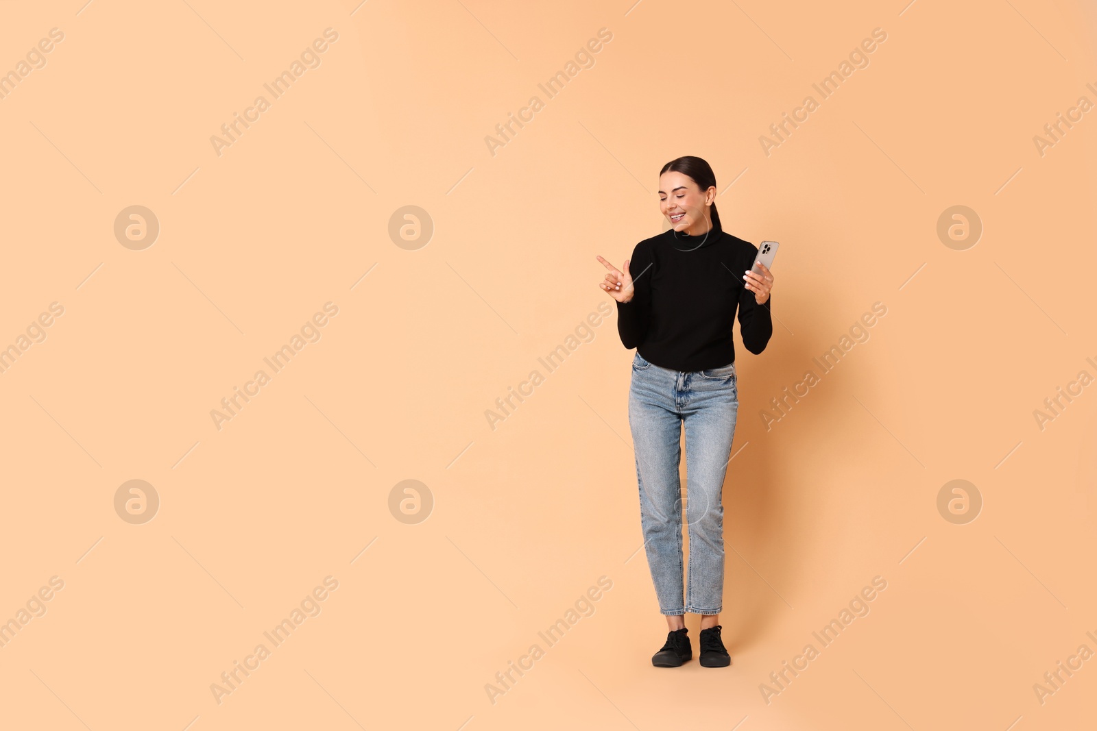 Photo of Smiling woman with smartphone pointing at something on beige background. Space for text