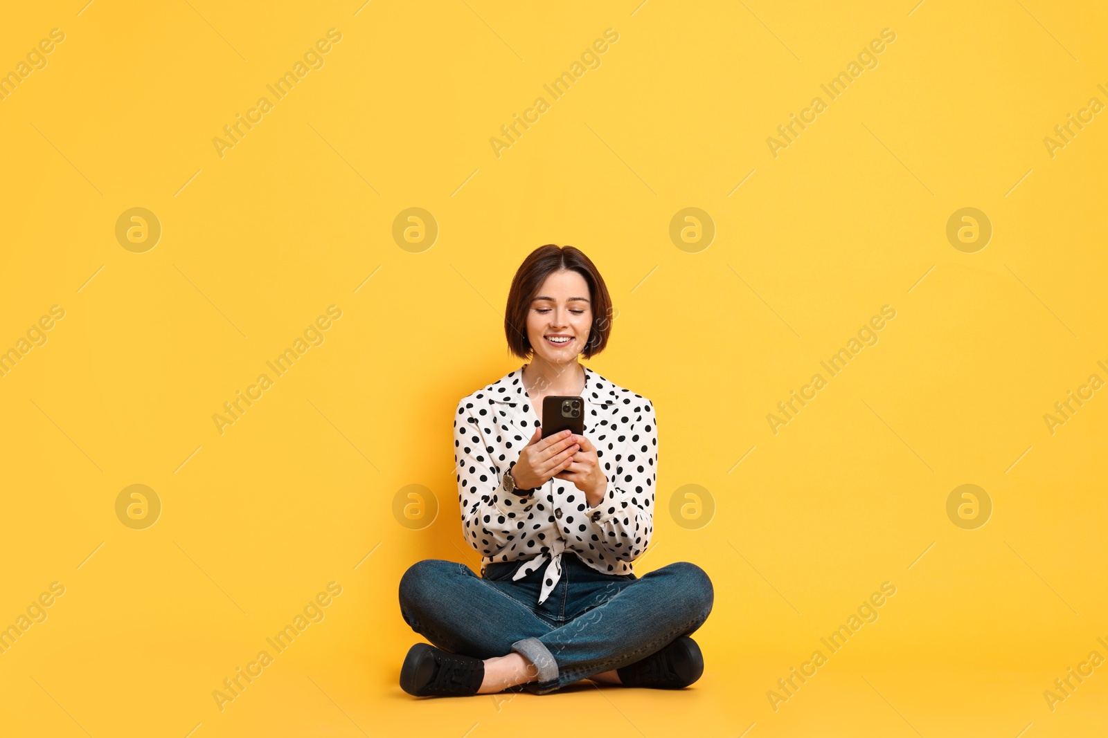 Photo of Smiling woman with smartphone sitting on yellow background