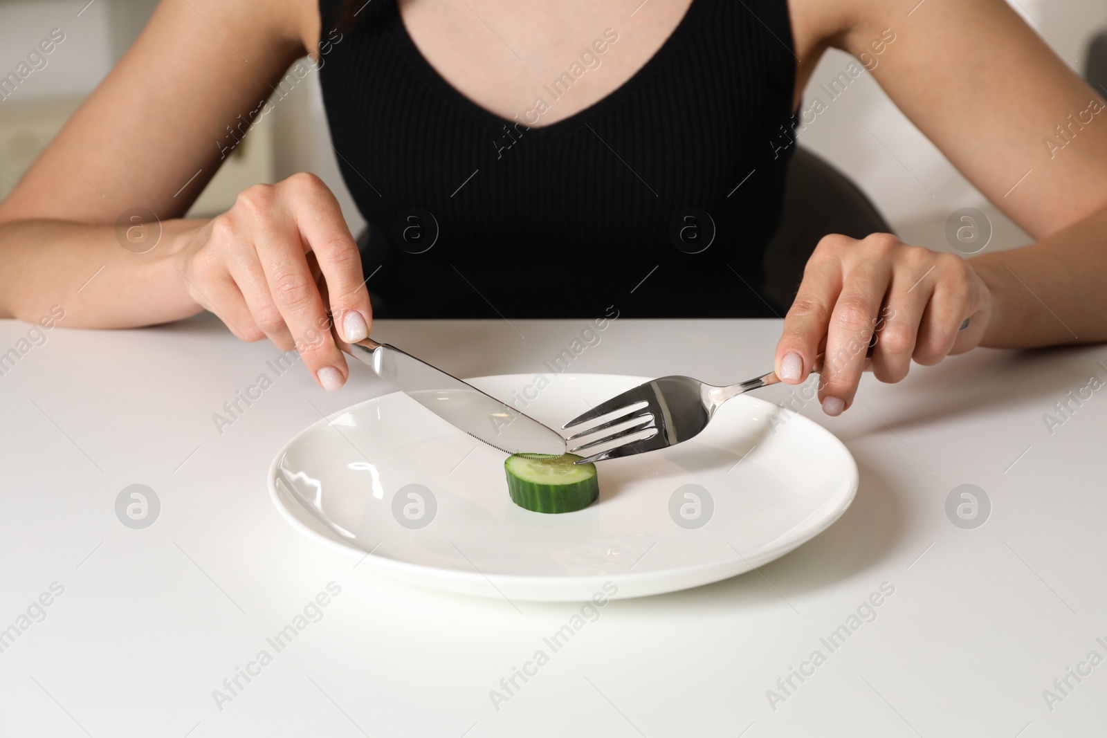 Photo of Eating disorder. Woman cutting cucumber at white table indoors, closeup