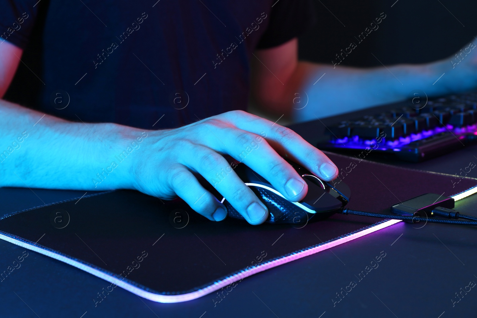 Photo of Gamer using modern wired computer mouse in neon lights at dark table, closeup