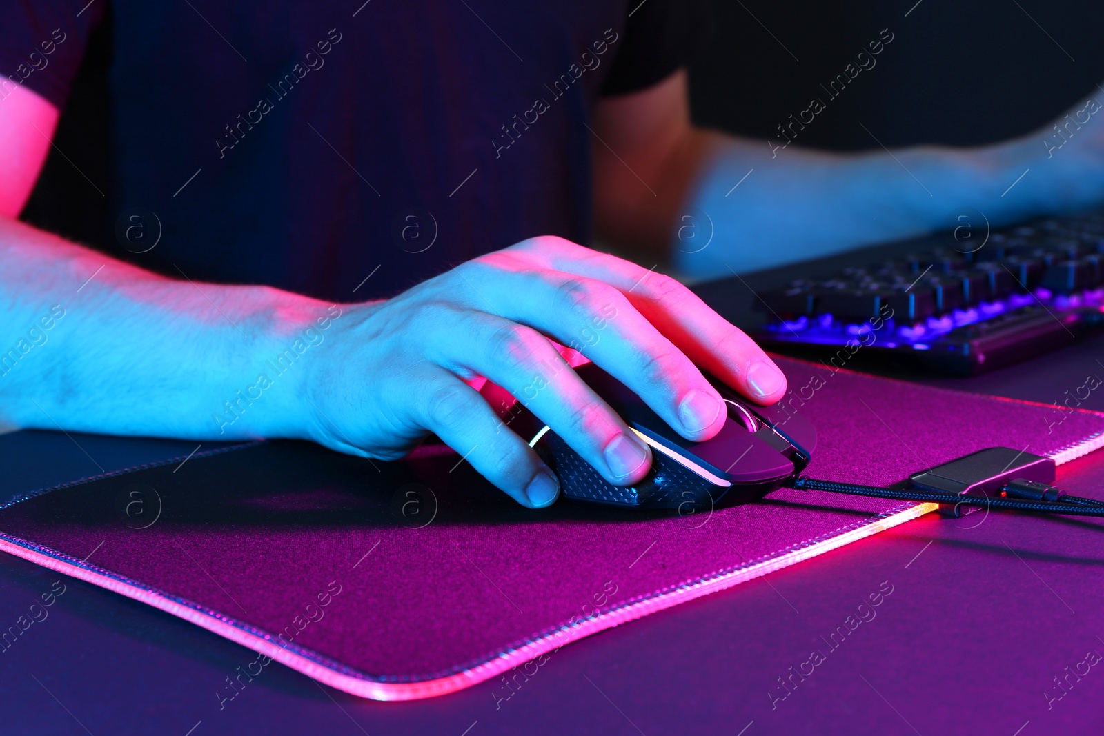 Photo of Gamer using modern wired computer mouse in neon lights at dark table, closeup