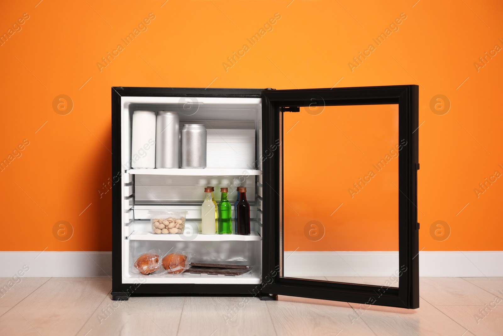 Photo of Mini refrigerator with different drinks and snacks near orange wall