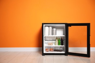 Mini refrigerator with different drinks and snacks near orange wall, space for text