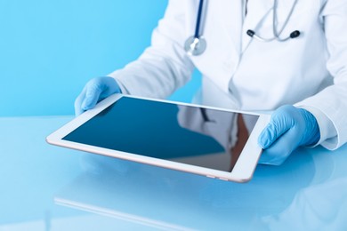 Doctor with tablet at table against light blue background, closeup view