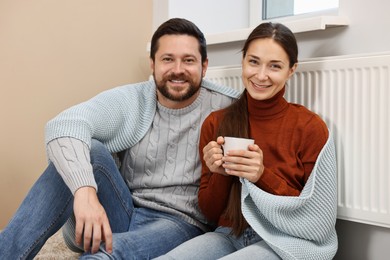 Happy couple with cup of hot drink near heating radiator at home