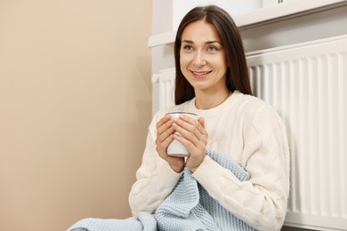 Photo of Happy woman with cup of hot drink near heating radiator at home