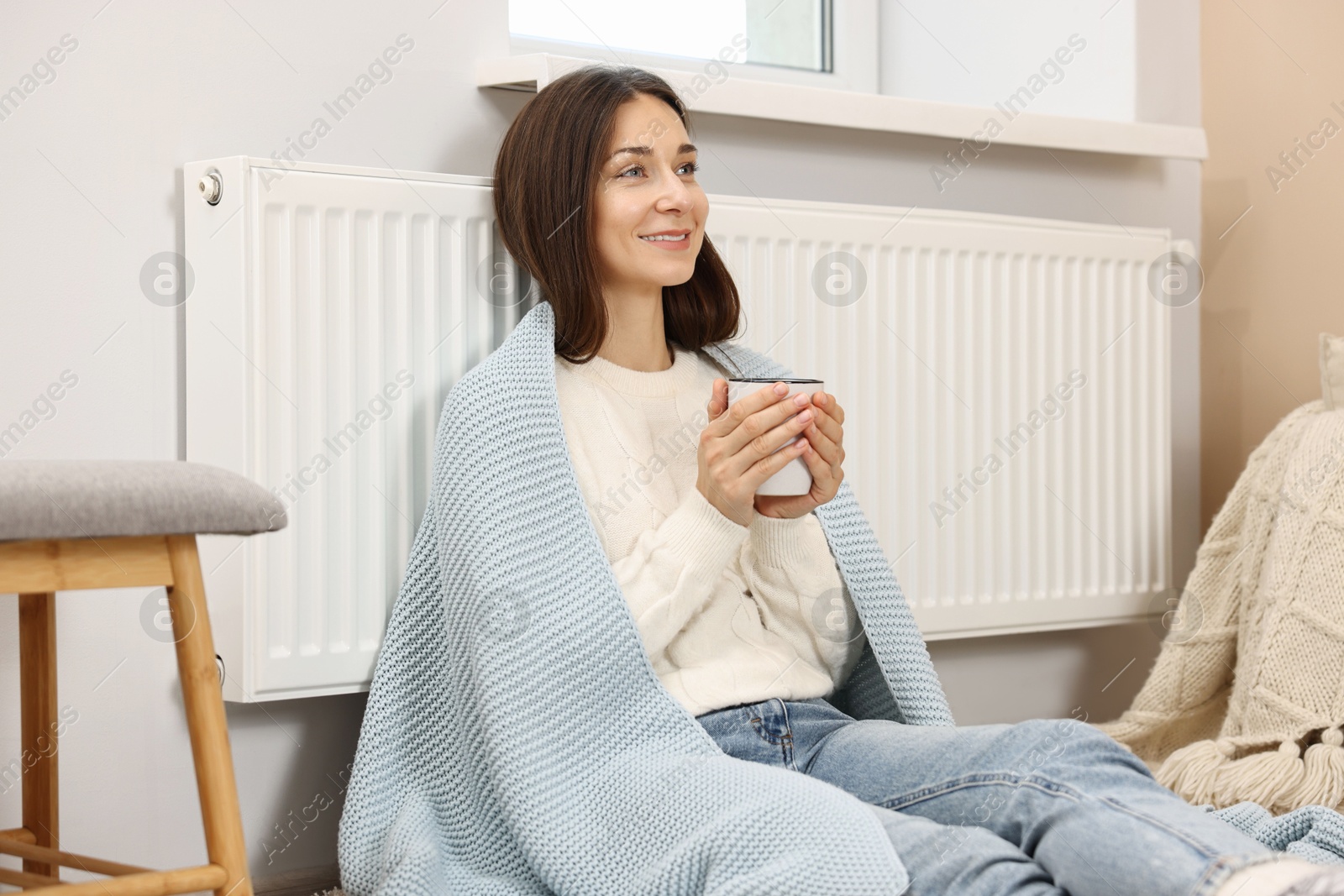 Photo of Happy woman with cup of hot drink near heating radiator at home