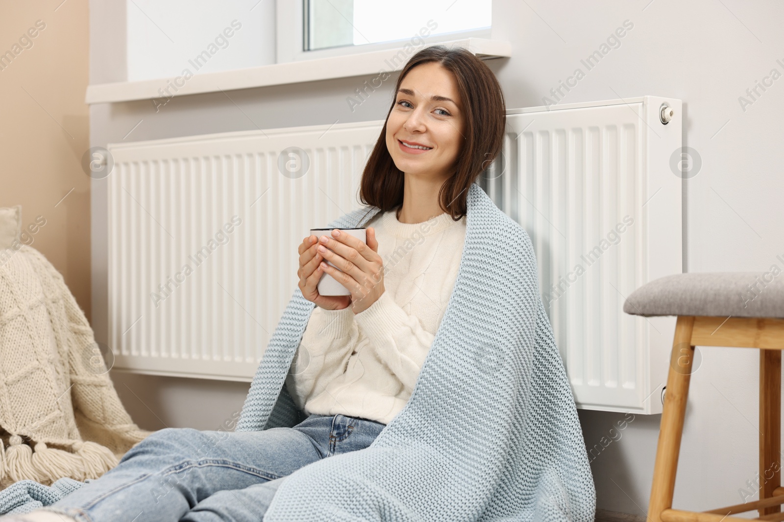 Photo of Happy woman with cup of hot drink near heating radiator at home