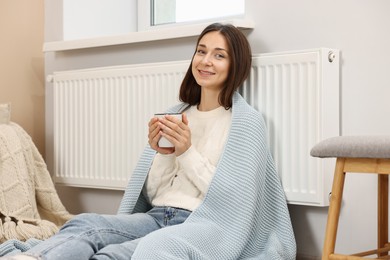 Happy woman with cup of hot drink near heating radiator at home