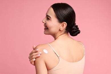 Photo of Smiling woman with cream on shoulder against pink background, back view. Body care