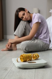 Eating disorder. Sad woman sitting near scale and measuring tape on floor indoors, selective focus