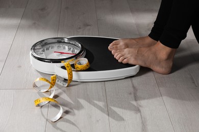 Photo of Eating disorder. Woman sitting near scale and measuring tape on floor indoors, closeup