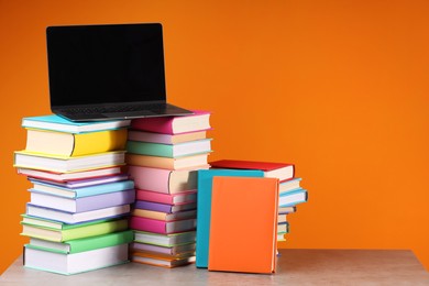 Stacks of colorful books and laptop on wooden table against orange background, space for text